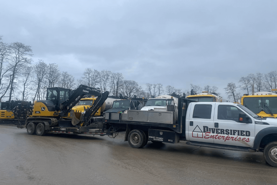 A white flatbed truck with "Diversified Enterprises" written on the side is parked on a muddy lot, hauling a trailer carrying a yellow John Deere mini excavator. Several other heavy-duty vehicles, including school buses and construction equipment, are visible in the background under an overcast sky.