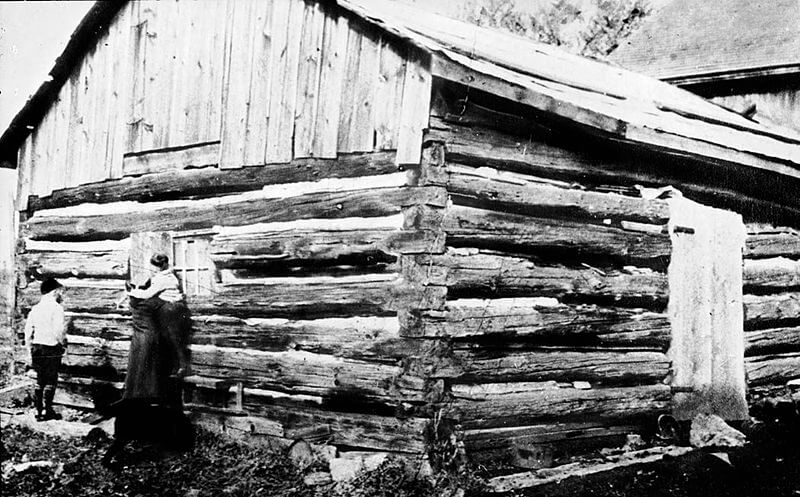 A black-and-white photograph of an old log cabin with a slanted wooden roof. A woman lifts a child to peek through a small window while a man stands nearby. The rough-hewn logs and simple construction suggest a historical rural setting.




