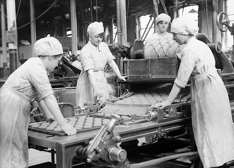 A black-and-white photo of women working in a factory, likely from the early 20th century. They wear aprons and caps while operating machinery that presses dough into uniform shapes.