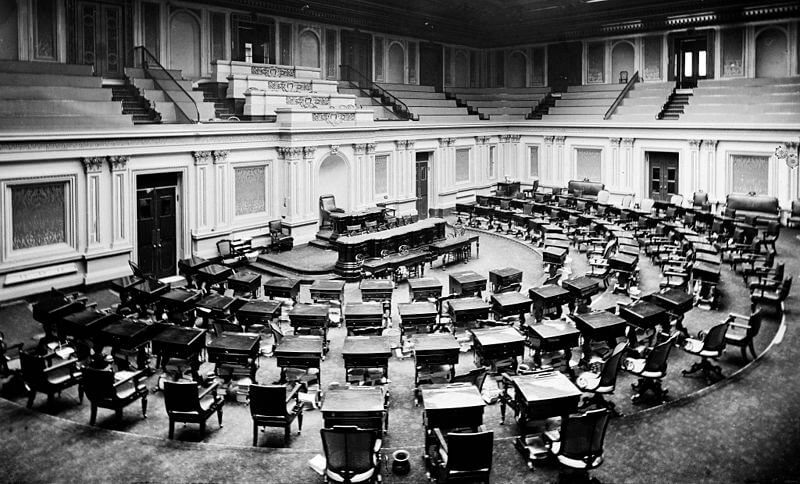 A black-and-white photograph of an empty legislative chamber with a semicircular arrangement of wooden desks and chairs. Ornate architectural details, high ceilings, and gallery seating surround the room, suggesting a historical government setting.