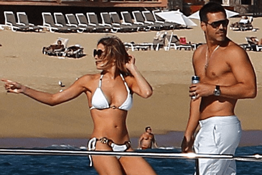 A woman in a white bikini and oversized sunglasses stands on a boat, gesturing and looking off to the side while adjusting her hair. Beside her, a shirtless man in white pants and sunglasses holds a beverage can, also gazing in the same direction. The background features a sandy beach with lounge chairs, umbrellas, and beachgoers.