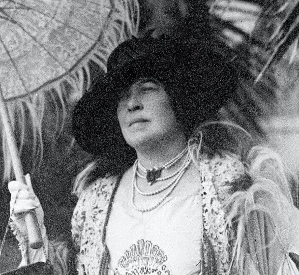 A black-and-white photograph of a woman dressed in elaborate early 20th-century fashion. She wears a large feathered hat, layered pearl necklaces, and a fur-trimmed coat while holding a decorative parasol. Her expression is poised and serious as she gazes into the distance.