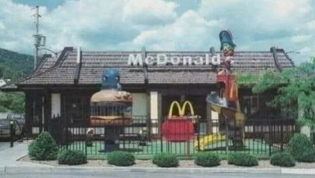 A retro McDonald's restaurant with a brown-tiled roof features a fenced-in playground. The playground includes a large Captain Crook slide, a Hamburglar jail climber, and a McDonald's-themed play structure with golden arches.