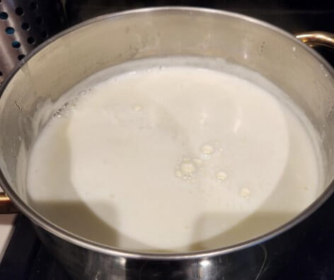 A stainless steel pot sits on the stovetop, filled with simmering milk. The surface is smooth and frothy, with small bubbles forming near the center.





