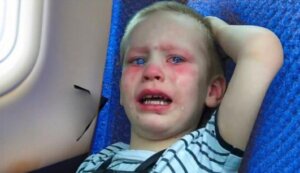 A young child with teary eyes and a distressed expression lies against the blue fabric of an airplane seat. Their flushed face and upset demeanor suggest discomfort or distress during the flight, with a window and part of the aircraft's interior visible nearby.