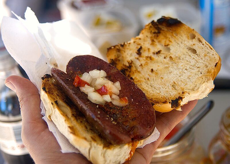 A hand holds a grilled sausage sandwich with a charred bun, slightly open to reveal the contents. The dark, seared sausage is topped with finely chopped onions and red peppers, adding a fresh contrast to the rich, smoky meat.