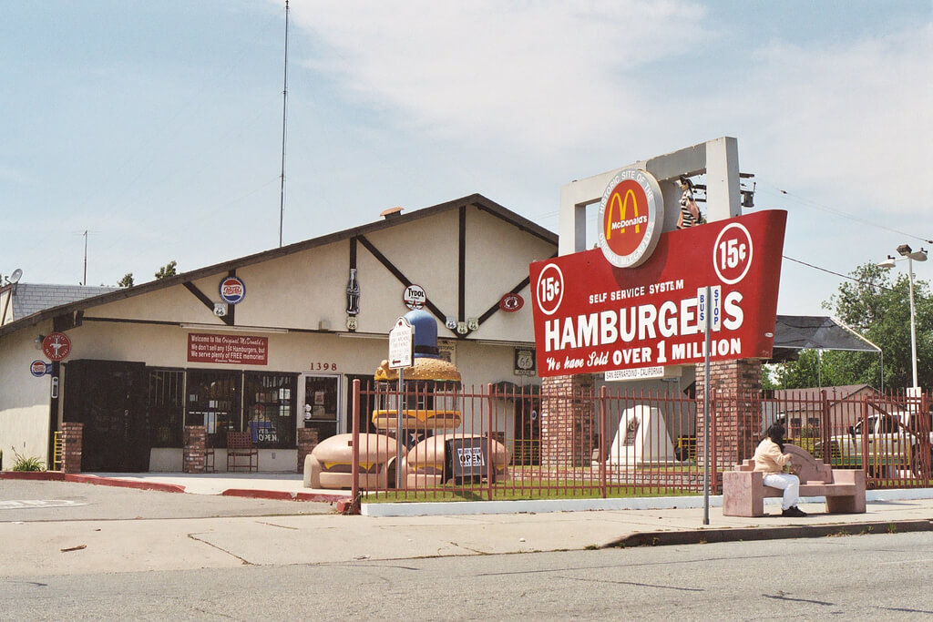 The original McDonald's site in San Bernardino, California, now a museum, features a vintage sign advertising 15-cent hamburgers. The building has nostalgic decor, oversized burger sculptures, and a fenced area, with a person sitting on a bench nearby.