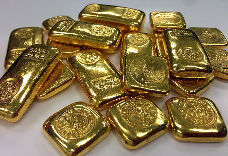 A close-up photograph of multiple gold bars stacked on a white surface. The bars are shiny, irregularly shaped, and embossed with markings, including weight and mint seals.