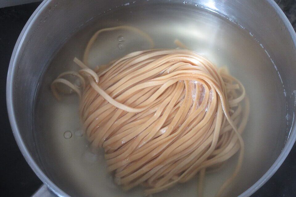 A bundle of uncooked noodles sits in a pot of hot water, beginning to soften and unravel. The water is slightly cloudy, and a few stray strands float around the tightly wound mass.