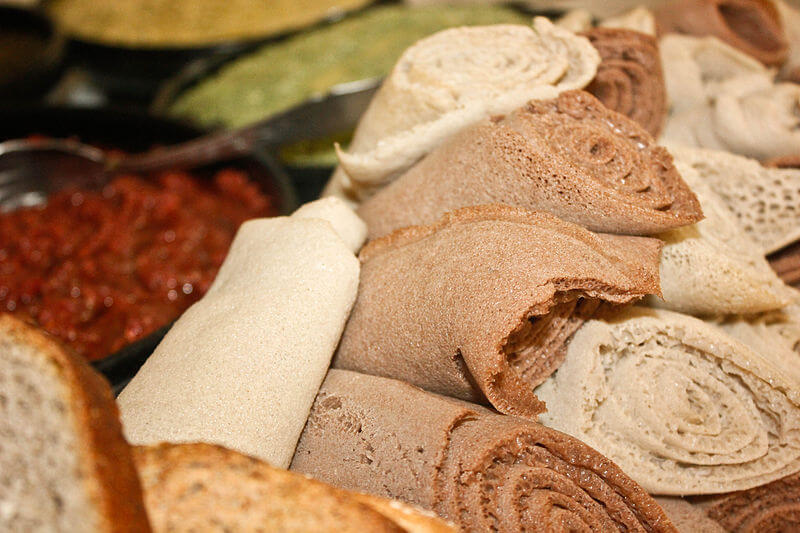 A plate holds rolled pieces of injera, the traditional Ethiopian flatbread, in shades of light beige and deep brown. In the background, bowls of rich red and green sauces add a colorful contrast to the spongy, porous texture of the bread.