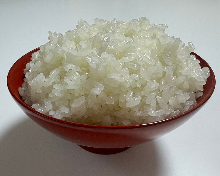 A red bowl filled with freshly cooked, glistening white rice sits on a plain white surface. The individual grains appear soft, slightly sticky, and slightly translucent, piled high above the bowl’s rim.