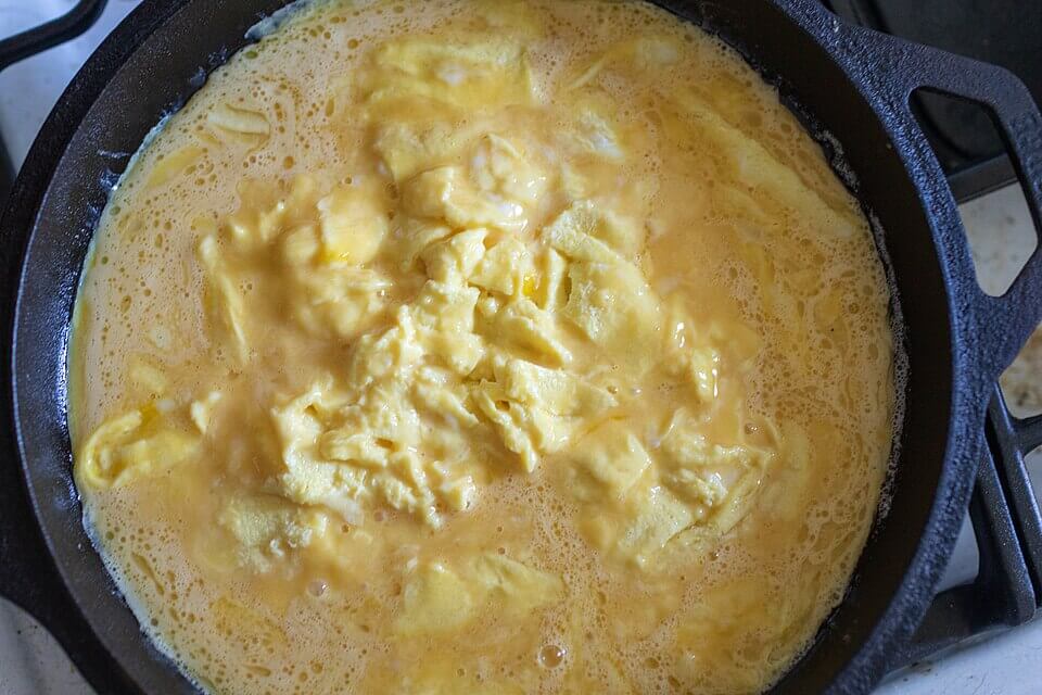 A cast iron skillet sits on a stovetop, filled with partially cooked scrambled eggs. The eggs are soft and slightly runny, with a creamy yellow texture and bubbling edges.