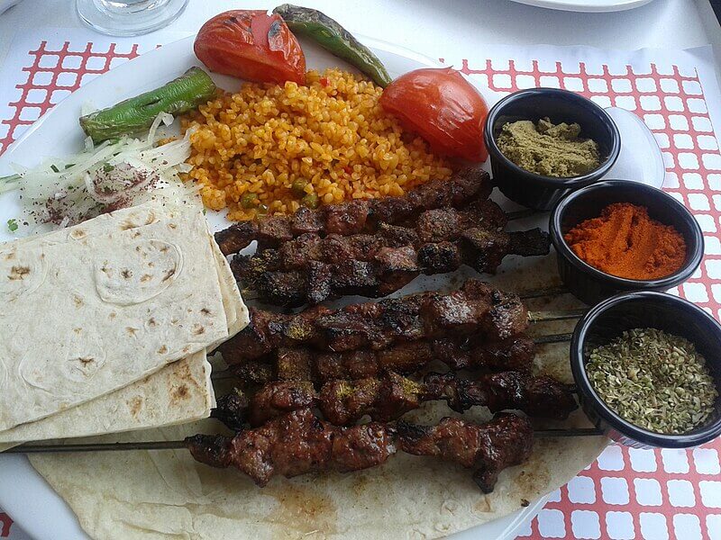 A plate of Turkish-style kebabs features skewered, char-grilled meat resting on flatbread. The meal is accompanied by bulgur pilaf, grilled vegetables, fresh onions, and small bowls of vibrant spices.