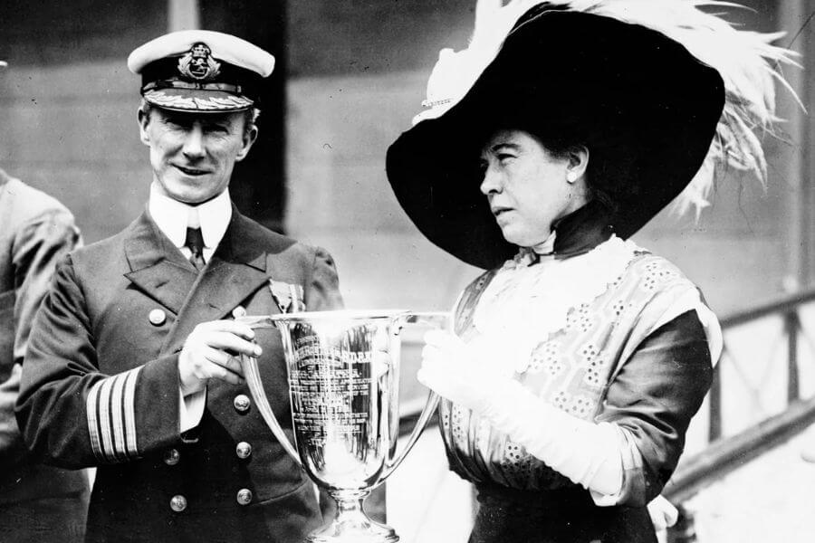 A black-and-white photograph of a naval officer and a woman holding a large trophy. The officer wears a decorated uniform and cap, while the woman is dressed in an elegant outfit with a wide-brimmed feathered hat and gloves. They both appear serious as they grip the trophy together.