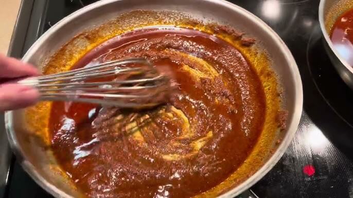 A hand uses a metal whisk to stir a rich, reddish-brown sauce in a pan on the stovetop. The thick mixture swirls smoothly, with flecks of spices visible along the edges of the pan.