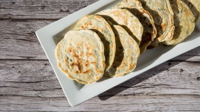 A white rectangular plate holds a neatly arranged stack of golden-brown scallion pancakes. The pancakes have crispy, slightly charred surfaces with flecks of green herbs visible throughout the dough.