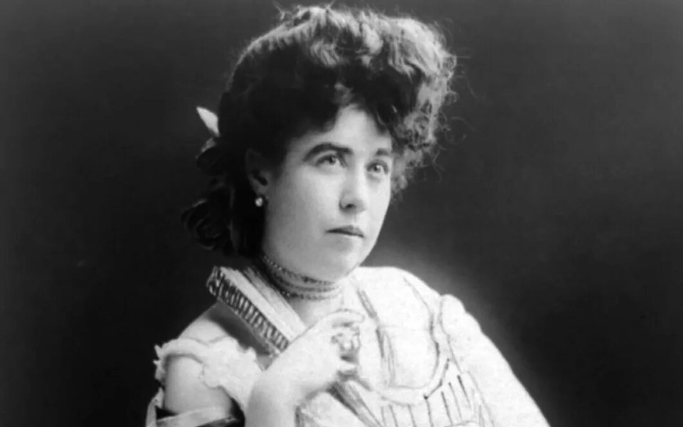 A black-and-white portrait of a woman from the late 19th or early 20th century. She has wavy hair, wears an elegant lace dress, and accessorizes with a choker and earrings. Her thoughtful gaze is directed upward, with one hand near her chin.