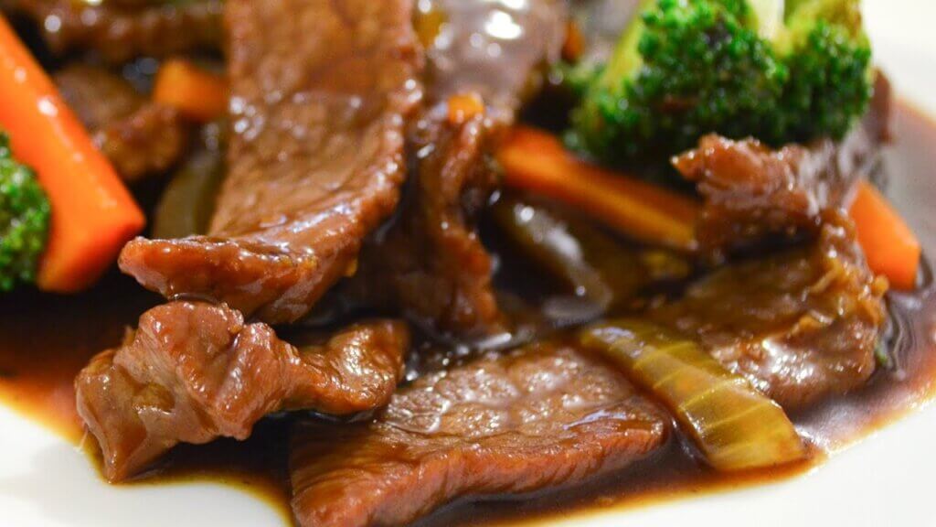 A plate of beef stir-fry features tender slices of glazed meat coated in a rich, glossy sauce. Bright green broccoli, crisp carrot sticks, and slivers of onion add vibrant color and texture to the dish.