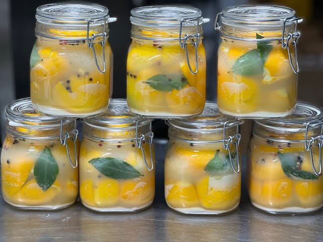 Six glass jars with metal clasps are neatly stacked, each filled with preserved yellow fruits submerged in brine. Floating bay leaves and whole peppercorns add a rustic, aromatic touch to the vibrant contents.