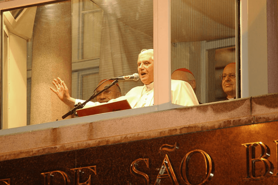 A Catholic cleric, likely a Pope, speaks into a microphone from a window balcony above a sign that reads "SÃO B...," with several cardinals standing beside him. He raises one hand mid-gesture while addressing a crowd outside at night.