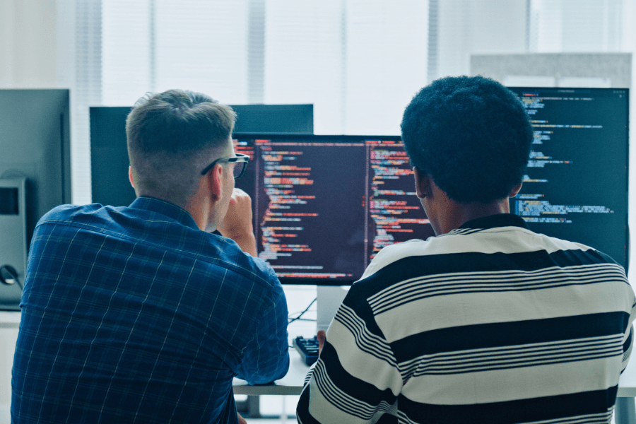 Two male programmers sit side by side in front of multiple monitors displaying colorful lines of code. They appear focused and deep in discussion, working together in a modern office environment.