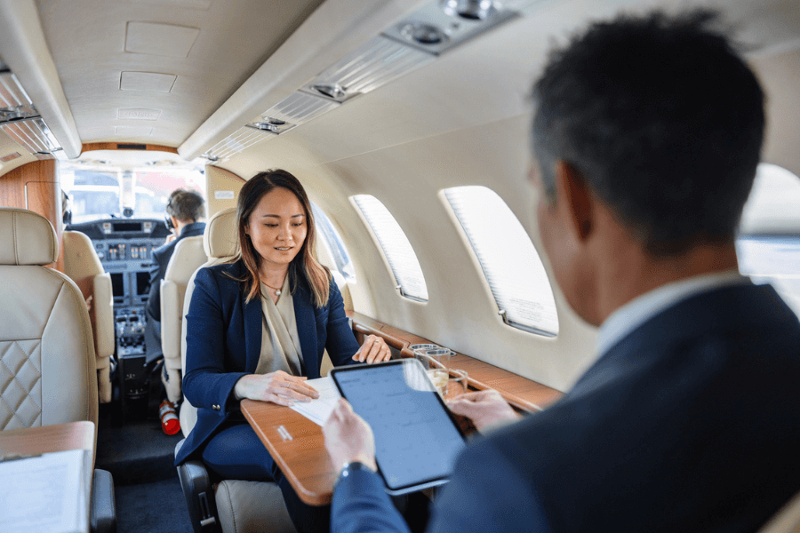 Two business professionals sit across from each other in a private jet, discussing documents and reviewing information on a tablet. The woman is dressed in a navy suit and appears to be explaining something, while the man listens attentively.