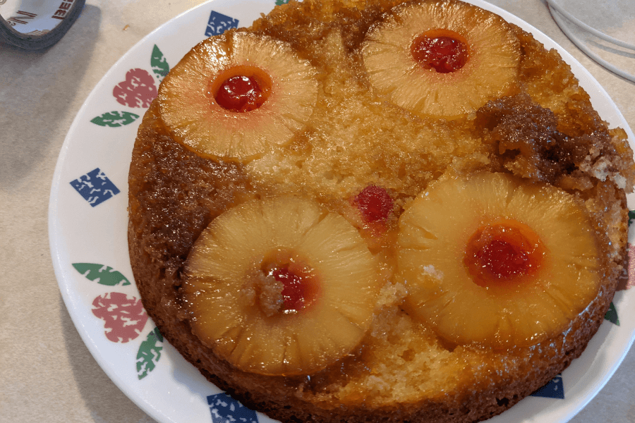 A pineapple upside-down cake on a floral-patterned plate, featuring caramelized pineapple rings and maraschino cherries baked into a golden brown, moist cake. A small section of the cake has been broken off at the edge.