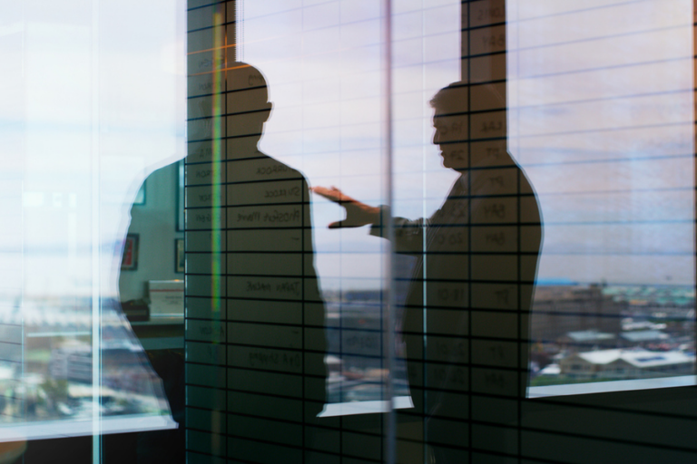 Silhouettes of two men having a discussion in an office, seen through a glass wall with a schedule or planning grid written on it. The reflection blends with the cityscape view outside, creating a layered and contemplative visual effect.