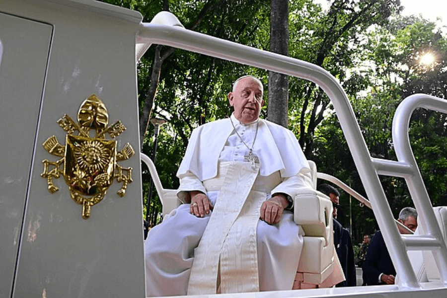The Pope sits in a white Popemobile surrounded by lush green trees, wearing his traditional white robes and cross pendant. A close-up of the Vatican coat of arms is visible on the vehicle, with sunlight filtering through the trees in the background.