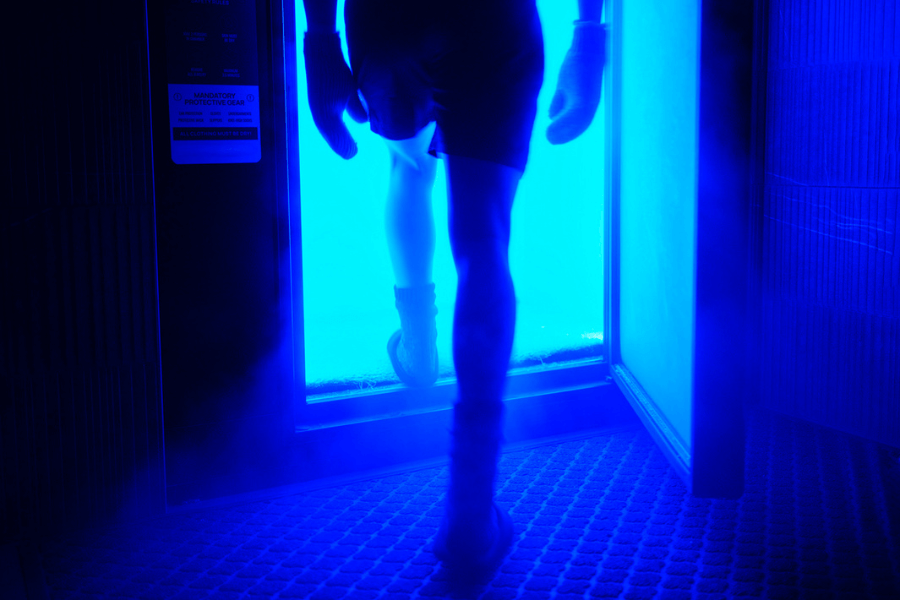 Person stepping into a cryotherapy chamber bathed in intense blue light, wearing protective gear including gloves, socks, and shorts. Cold vapor surrounds the entrance, emphasizing the futuristic and chilling environment.