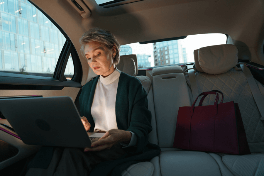 Elegant older woman working on a laptop in the backseat of a luxury car, with a red handbag placed beside her. She appears focused and composed, with city buildings visible through the window in the background.