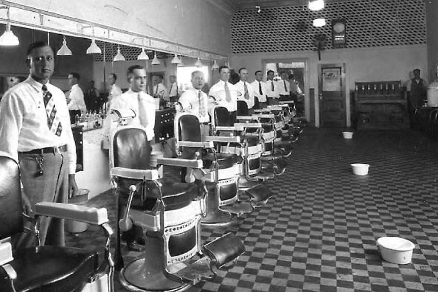 Black and white photo of a vintage barbershop with a row of classic barber chairs and uniformed barbers standing beside them. The shop features checkered flooring, wall mirrors, and overhead pendant lights, evoking a mid-20th century setting.