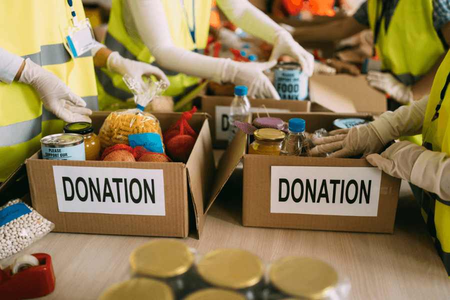 Volunteers wearing yellow safety vests and gloves pack cardboard boxes labeled “DONATION” with non-perishable food items, including canned goods, pasta, beans, and bottled water. The setting suggests a community food drive or charity event.
