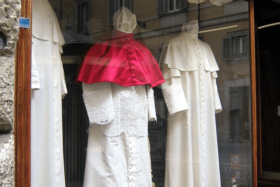 Three mannequins dressed in traditional clerical garments are displayed in a storefront window. The center outfit features a white cassock with lace and a bright red shoulder cape, while the other two wear simpler white cassocks with shoulder capes and zucchettos, reflecting Catholic clerical attire.