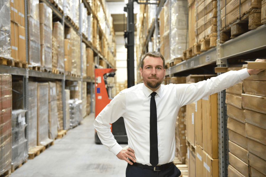 Confident man in a white shirt and black tie stands in a large warehouse aisle filled with stacked boxes and pallets, with a forklift visible in the background. He poses with one hand on his hip and the other resting on a shelf.