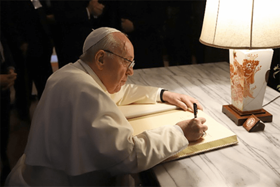 The Pope, dressed in white papal robes and zucchetto, signs a guestbook at a marble desk under the warm light of a floral-patterned table lamp. The atmosphere is formal and respectful, with people standing in the background.