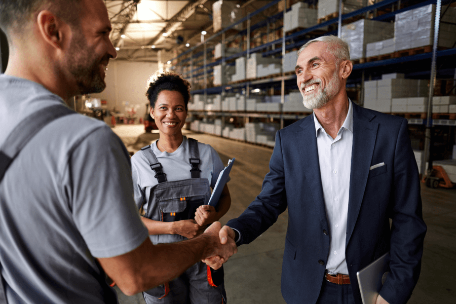 Smiling businessman in a navy suit shakes hands with a warehouse worker in grey overalls, while another worker holding a clipboard watches with a cheerful expression. They are standing in a spacious warehouse with shelves stocked with boxes.