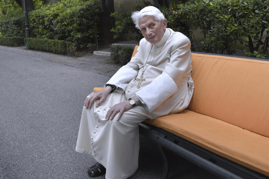 An elderly Catholic clergyman dressed in white robes and wearing a cross necklace sits on an orange-cushioned bench in a peaceful outdoor setting. He looks gently toward the camera, surrounded by greenery on a paved path.