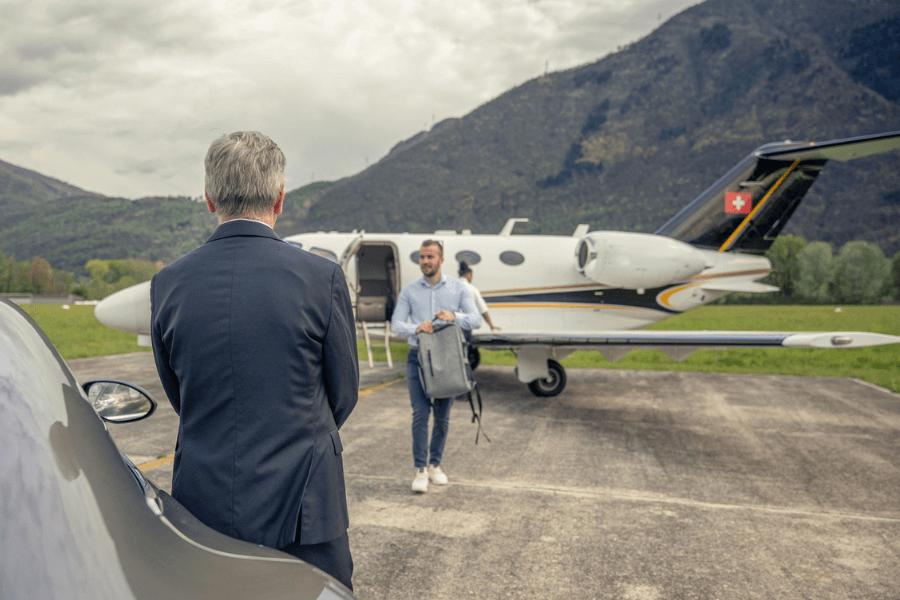 A man in a business suit stands next to a luxury car on a private airstrip, facing another man walking toward him from a small private jet with a Swiss flag on the tail. The arriving man is casually dressed, carrying a grey backpack, with mountains and cloudy skies in the background.