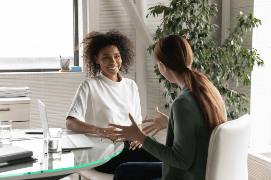 Two women engaged in a friendly conversation in a bright office setting, sitting at a glass desk with a laptop and water glasses. One woman is smiling while the other gestures as she speaks, indicating an engaging and collaborative discussion.