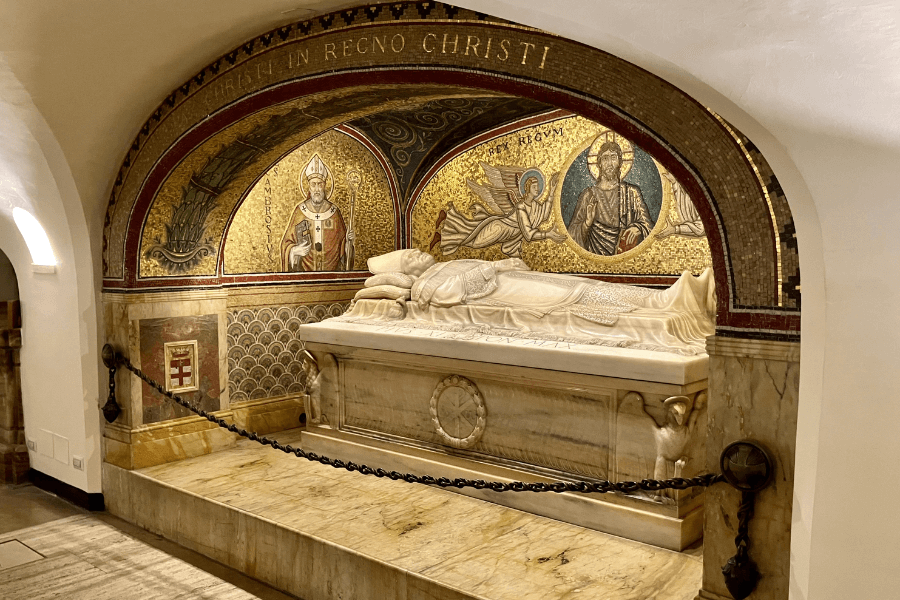 An ornate marble tomb with a reclining effigy of a cleric lies beneath a semicircular mosaic featuring religious figures and Latin inscriptions. The artwork includes depictions of Saint Ambrose and Christ, with the words "CHRISTI IN REGNO CHRISTI" above, within a chapel or crypt setting.