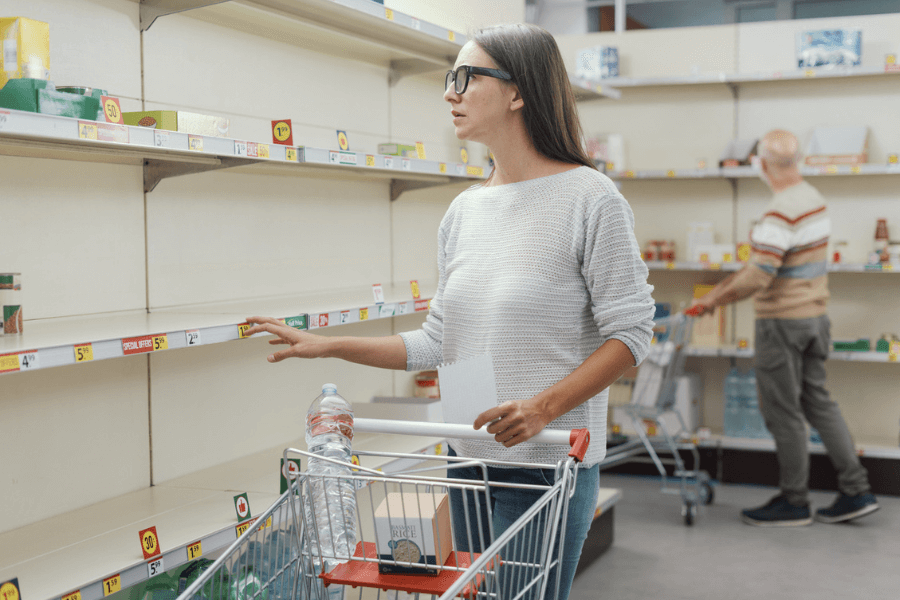 Woman holding a shopping list stands in front of nearly empty supermarket shelves, looking concerned while placing items in her cart. Another shopper is seen in the background, also browsing the sparse inventory.