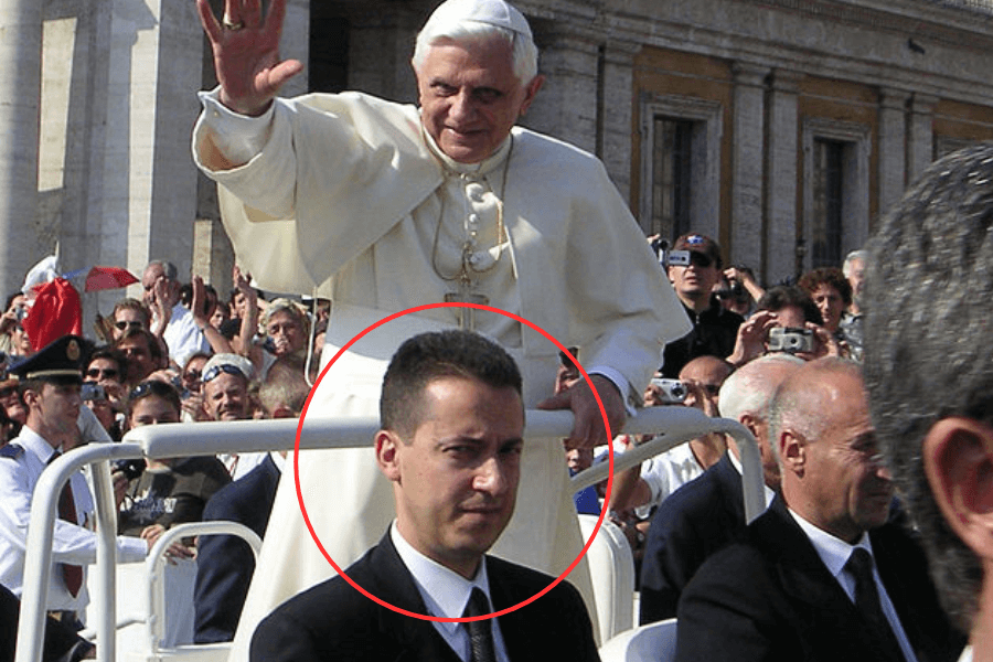 Pope Benedict XVI waves to a crowd while standing in the Popemobile during a public appearance. A man in a dark suit, circled in red, sits in front of him with a serious expression, surrounded by security personnel and onlookers.