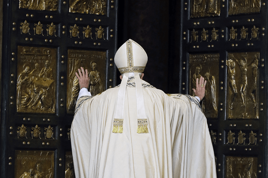 A pope, seen from behind, dressed in cream-colored liturgical vestments and a mitre, places his hands on the ornate bronze Holy Door adorned with religious reliefs at St. Peter’s Basilica. The door is partially open, symbolizing a significant ceremonial event.