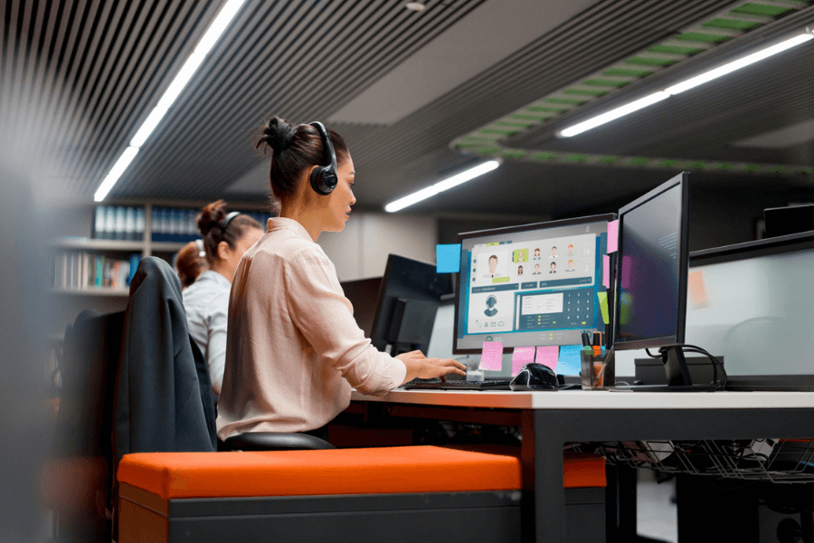 Woman wearing a headset works at a dual-monitor setup in a modern office, possibly as part of a customer support or call center team. She is focused on her screen, which displays a user interface with contact profiles and communication tools.