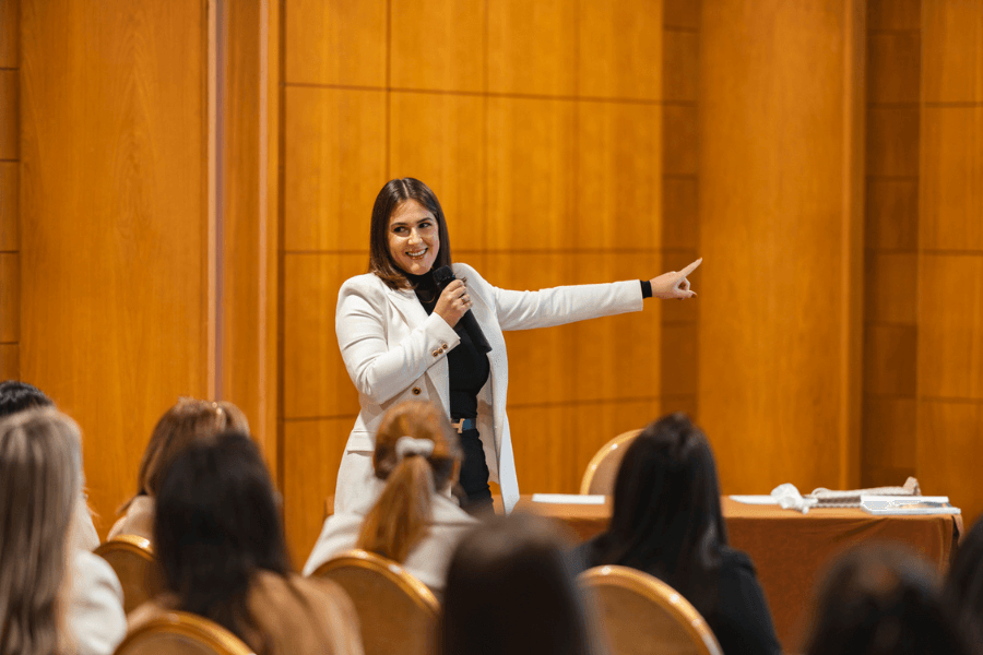 Confident woman in a white blazer holds a microphone and gestures while speaking to an audience in a formal setting with wood-paneled walls. Attendees, mostly women, listen attentively, suggesting a seminar, workshop, or conference.