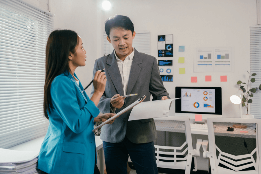 Two professionals in a modern office discuss data while holding clipboards, standing near a desk with a large computer monitor displaying charts and graphs. The workspace is organized with charts on the wall and colorful sticky notes.