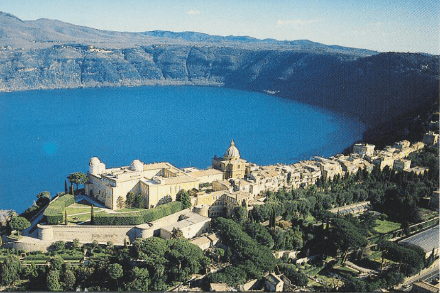 Aerial view of the papal summer residence, Castel Gandolfo, perched on the edge of the deep blue Lake Albano and surrounded by lush gardens and the town’s historic buildings. The scenic volcanic crater lake is encircled by rolling hills in the background.