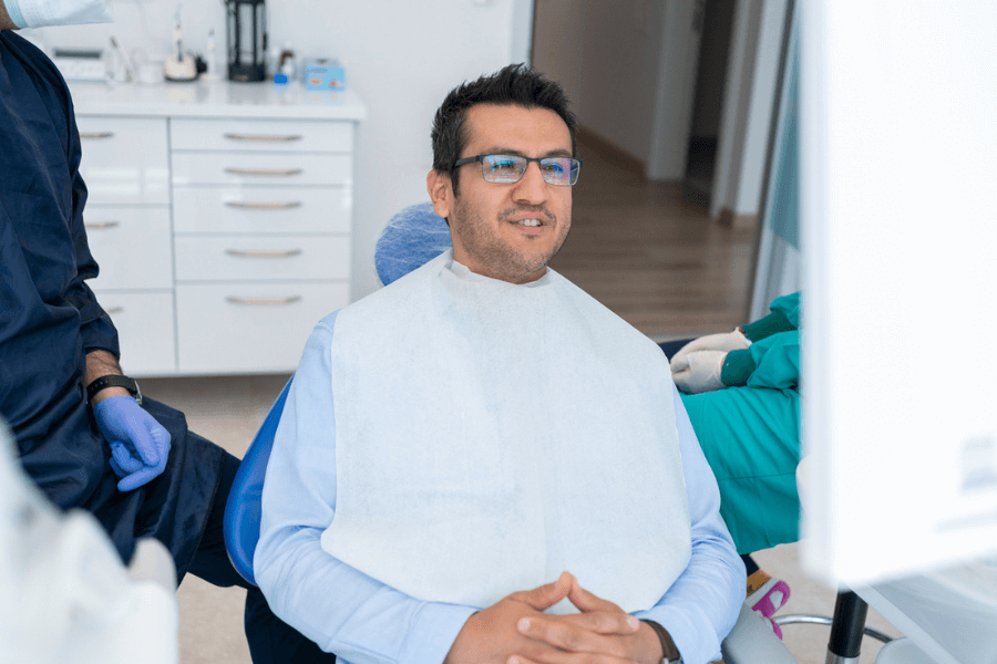 Man wearing glasses sits in a dental chair with a protective bib, smiling slightly as he interacts with the dentist, whose gloved hands are visible. The dental clinic is bright and modern with white cabinetry and clean surfaces.