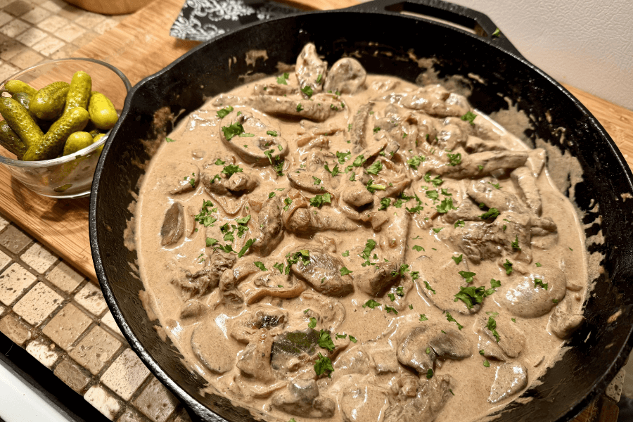 A cast iron skillet filled with beef stroganoff, featuring tender beef strips and mushrooms in a creamy brown sauce, garnished with chopped parsley. The skillet sits on a wooden cutting board next to a small glass bowl of pickles.